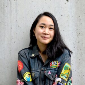 Headshot of a woman wearing a denim jacket covered in patches, leaning against a concrete wall