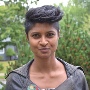 A headshot of Ninotchka Sequeira, smiling at the camera. She is wearing a leather jacket and standing outside, surrounded by plants. She has short hair that's cropped on the sides. 