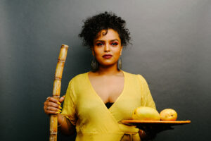 A woman with dark curly hair stands in the centre of a dark gray background. She is wearing large circular earrings, and a bright yellow dress. In her lefthand she is holding a stick, and in her right hand is a plate with yellow fruit. 