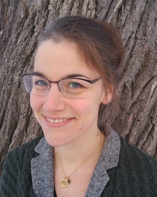 A headshot of Rachel Nolan, standing in front of a tree and smiling at the camera. She is wearing glasses, has light brown hair, and a gray collared shirt. 