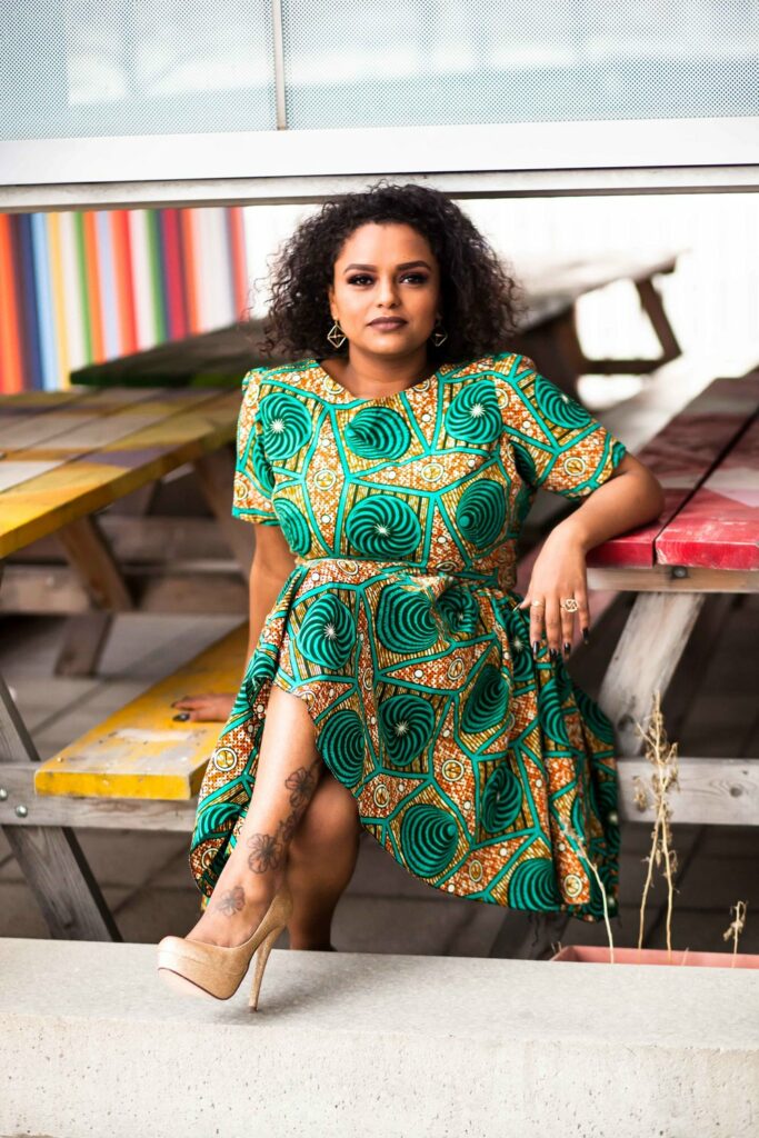 A woman with dark curly hair sits on a bench. She's wearing a greenand gold dress and heels. Behind her are shelves of multi-coloured books.