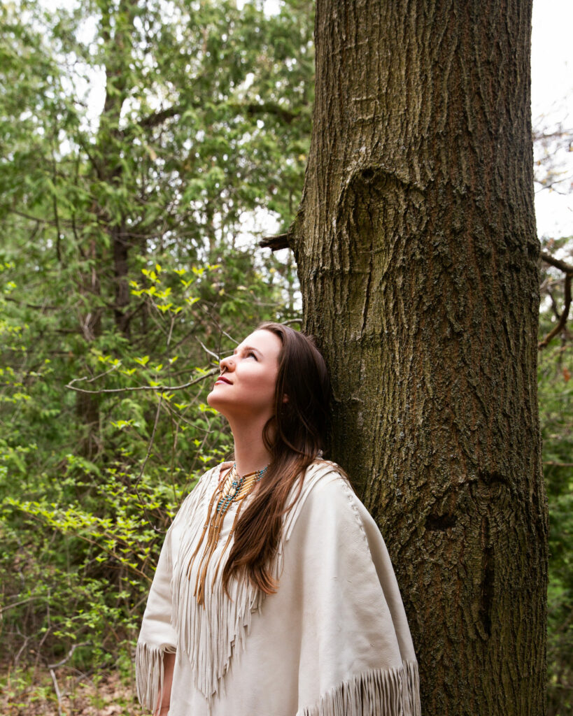 A woman in a tan shirt with fringe leans against a tree in the woods, looking upwards. She has long light brown hair and a septum piercing and nose ring.