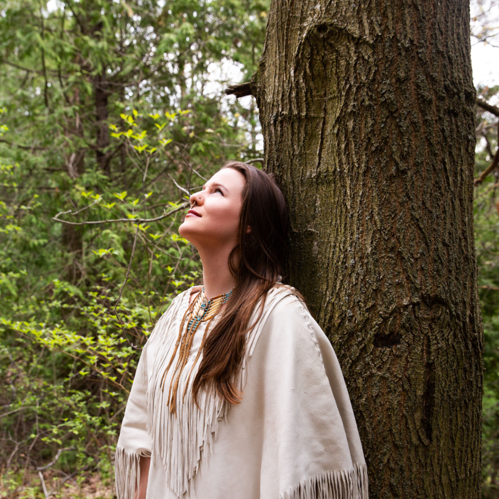 A woman in a tan shirt with fringe leans against a tree in the woods, looking upwards. She has long light brown hair and a septum piercing and nose ring.