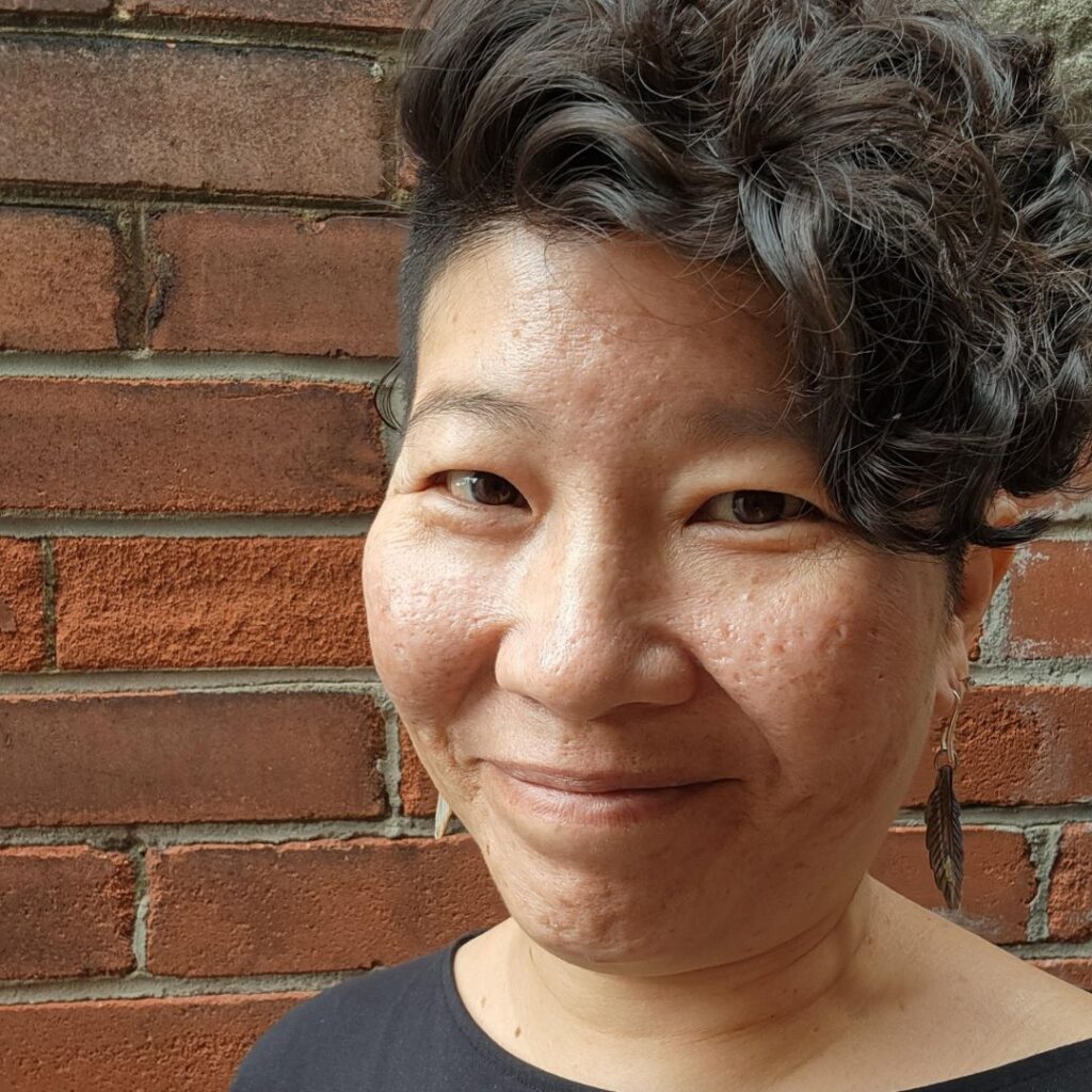 Headshot of Bo Yih -short, curly dark hair, smiling at the camera, and standing in front of a brick wall.