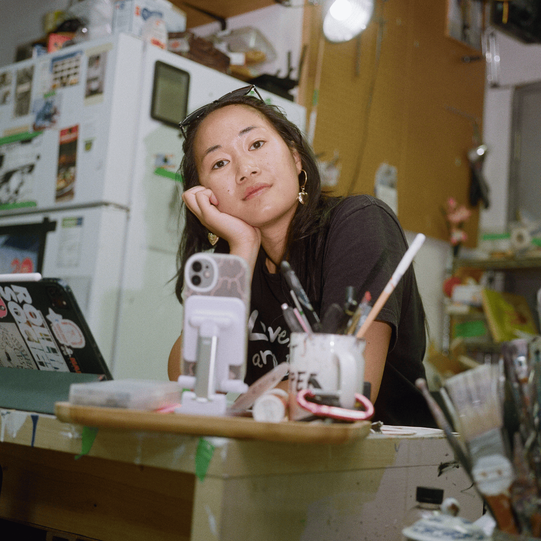 A woman leaning on the palm of her hand while sitting at a desk in an art studio. She has long dark hair, there is an open laptop next to her.